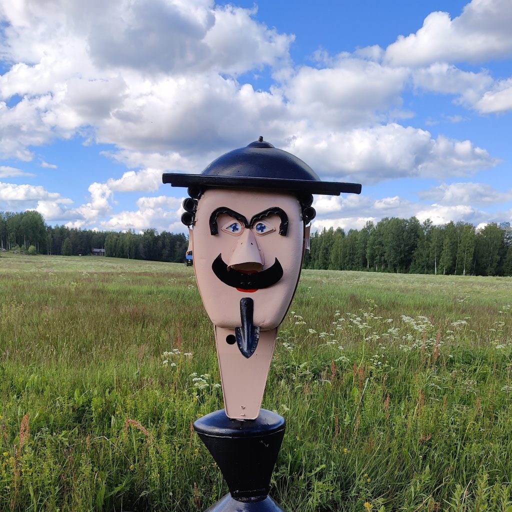 Vesa Väänäsen mustahattuinen pääfiguuri heinäpeltomaisemassa