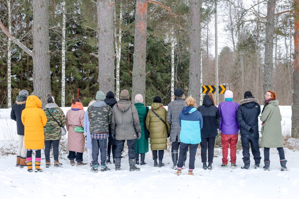 Yli 10 ihmistä seisoo rivissä selin kameraan päin talvisessa maisemassa. Taustalla isoja puita. Oikeassa reunassa oleva henkilö muista poiketen katsoo kameraan päin ja hymyilee