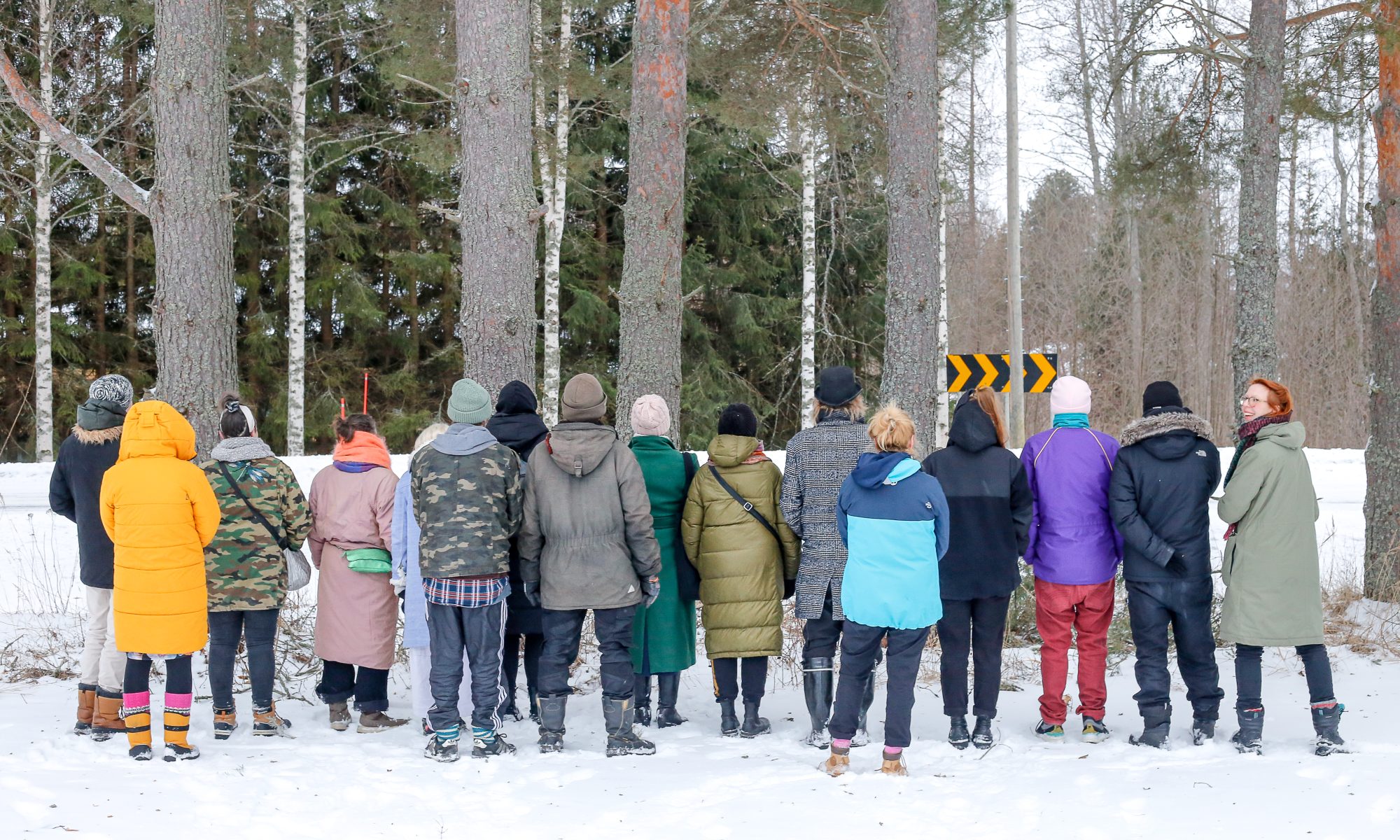 Yli 10 ihmistä seisoo rivissä selin kameraan päin talvisessa maisemassa. Taustalla isoja puita. Oikeassa reunassa oleva henkilö muista poiketen katsoo kameraan päin ja hymyilee