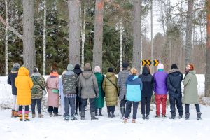 Yli 10 ihmistä seisoo rivissä selin kameraan päin talvisessa maisemassa. Taustalla isoja puita. Oikeassa reunassa oleva henkilö muista poiketen katsoo kameraan päin ja hymyilee